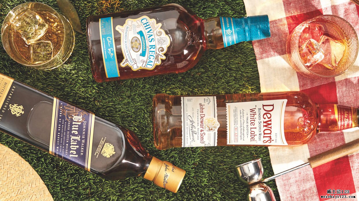 一瓶瓶混合苏格兰威士忌——尊尼获加蓝牌、杜瓦的白牌和芝华士水楢——从鸟瞰图上看，放在它们的两侧，上面似乎是短草，旁边是红白格子——带图案的野餐毯，还有一个木柄金属搅拌器，以及两个装有冰块和威士忌鸡尾酒的玻璃杯； 从照片的左下角可以看到草帽的边缘部分。